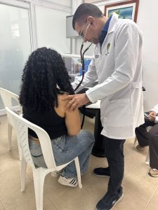 Un médico con bata blanca ausculta la espalda de una paciente sentada en una silla plástica blanca dentro de un consultorio.