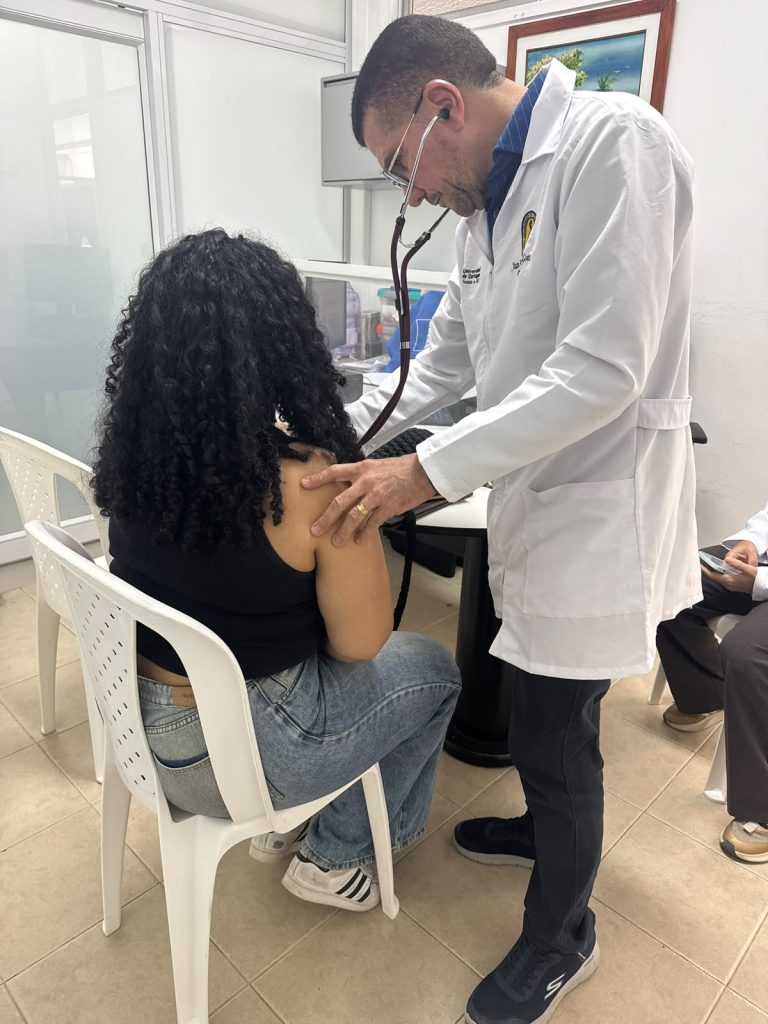 Un médico con bata blanca ausculta la espalda de una paciente sentada en una silla plástica blanca dentro de un consultorio.