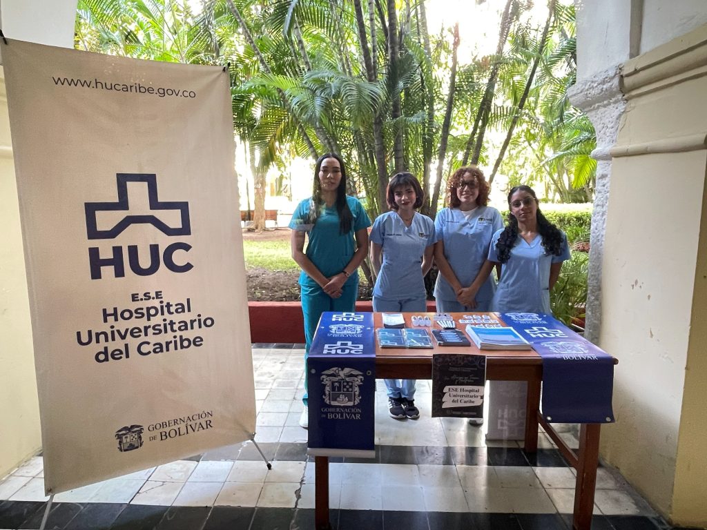 Cuatro estudiantes de Trabajo Social vistiendo uniforme médico al lado de un pendón del Hospital Universitario del Caribe y una mesa de información institucional con folletos de atención humanizada.