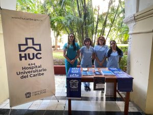 Cuatro estudiantes de Trabajo Social vistiendo uniforme médico al lado de un pendón del Hospital Universitario del Caribe y una mesa de información institucional con folletos de atención humanizada.