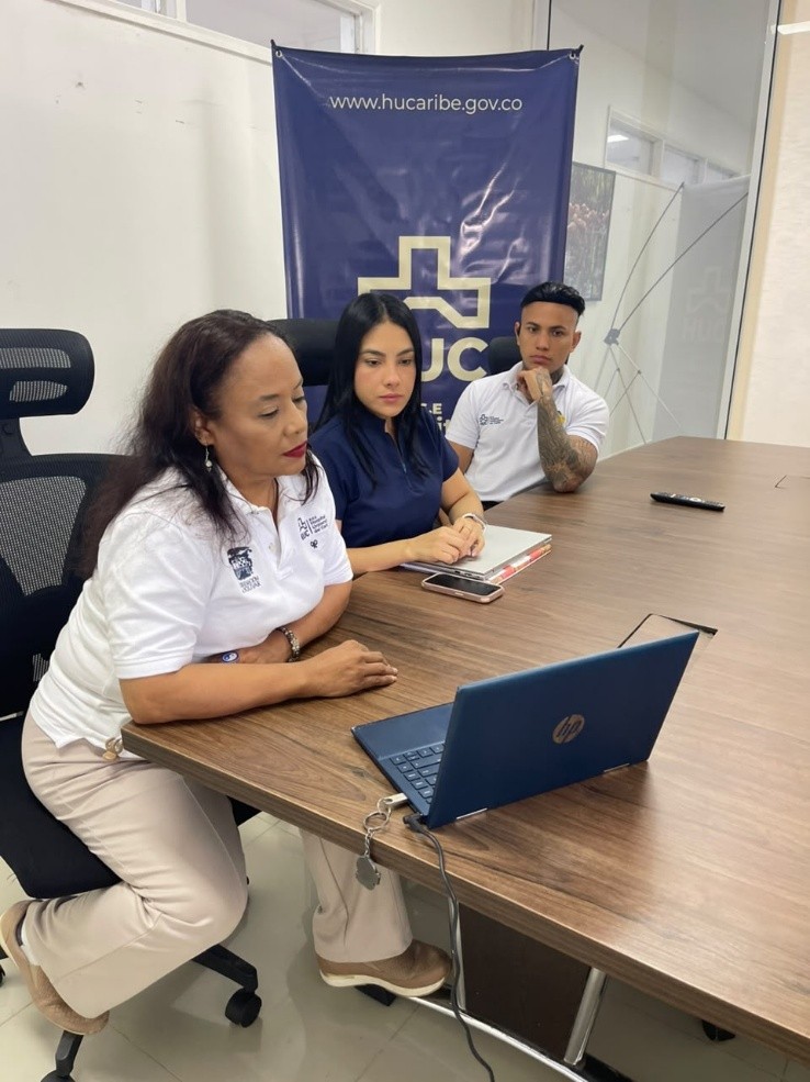 Tres colaboradores del Hospital Universitario del Caribe sentados frente a un computador portátil en una mesa de reuniones durante una sesión de trabajo o capacitación institucional, con un pendón del HUC de fondo.